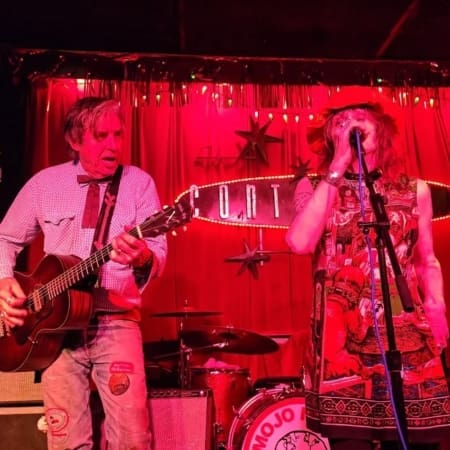 A man playing guitar and a woman singing perform on stage under red lighting, with a drum set and a "MOJO" sign in the background. Both appear deeply engaged—capturing the energy of live music Austin is famous for. Austin Marathon Half Marathon & 5K