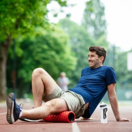 A man in a blue shirt and shorts smiles while foam rolling his thigh on a running track outdoors, preparing for marathon success. A water bottle rests beside him, with trees visible in the background. Austin Marathon Half Marathon & 5K
