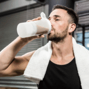 A man with a beard and short hair, wearing a black tank top and a white towel around his neck, drinks from a protein shaker bottle in a gym setting—highlighting what runners should eat post-workout. Austin Marathon Half Marathon & 5K