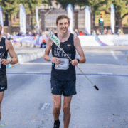 A male runner wearing a race bib and black athletic gear crosses a marathon finish line holding a pace sign, smiling, after expertly guiding others at the ideal marathon pace, with spectators and banners visible in the background. Austin Marathon Half Marathon & 5K