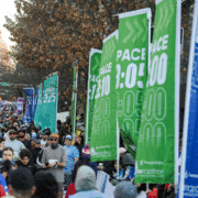 A large crowd of marathon runners fills a city street, moving past tall green banners displaying ideal marathon paces like “PACE 3:05” and “PACE 3:10,” with autumn trees and a building dome in the background. Austin Marathon Half Marathon & 5K