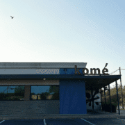 A modern restaurant building in Austin with a sign reading "komé" on the roof, blue tiles on the wall, large windows, and a clear sky with a single bird flying overhead—a hidden gem perfect for post-run recovery. Austin Marathon Half Marathon & 5K