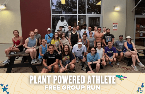 A diverse group of smiling runners poses outside a building, some sitting on picnic benches and others standing behind. A banner at the bottom reads "Plant Powered Athlete Free Group Run," highlighting this inclusive group run event. Austin Marathon Half Marathon & 5K