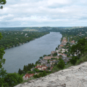 View from a rocky hilltop overlooking a wide river lined with green trees and houses; the river curves through a residential area with cloudy skies above, making it one of the most scenic runs among local running routes in Austin. Austin Marathon Half Marathon & 5K