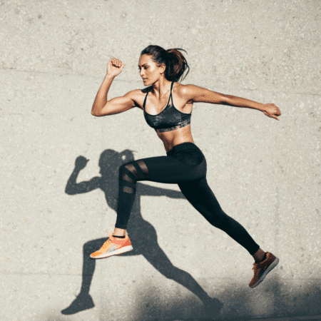 Athletic woman in sportswear leaps in midair against a concrete wall, casting a strong shadow. Dressed for running training, she wears a black sports bra, black leggings, and orange sneakers, looking focused and energetic. Austin Marathon Half Marathon & 5K