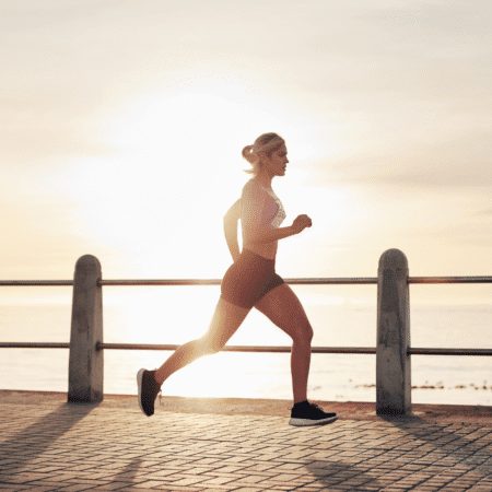 A woman jogs along a seaside promenade at sunrise, the sun low behind her. Dressed in athletic clothing, she appears focused on her running training as she runs near the water. Austin Marathon Half Marathon & 5K
