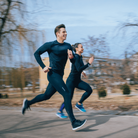 Two people in dark athletic wear are jogging side by side outdoors on a paved path, trees and buildings blurred in the background, suggesting fast motion—perfect for a tempo runs session during their running training. Austin Marathon Half Marathon & 5K