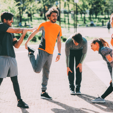 A group of five adults in athletic wear stretches together outdoors on a sunny day in a park, practicing injury prevention strategies as they prepare for exercise. Trees and greenery are visible in the background. Austin Marathon Half Marathon & 5K