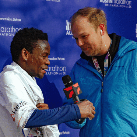 A marathon runner wrapped in a white blanket smiles and shakes hands with a reporter in a blue jacket holding a microphone, as photography captures the post-race interview in front of a blue event backdrop. Austin Marathon Half Marathon & 5K A marathon runner wrapped in a white blanket smiles and shakes hands with a reporter in a blue jacket holding a microphone, as photography captures the post-race interview in front of a blue event backdrop. Austin Marathon Half Marathon & 5K