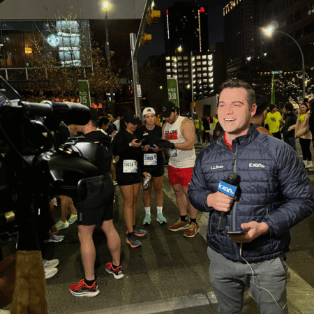 A reporter holding a microphone smiles at the camera on a city street at night, covering runners in race gear as a cameraman films for media requests. City buildings glow in the background, capturing dynamic photography of the event. Austin Marathon Half Marathon & 5K A reporter holding a microphone smiles at the camera on a city street at night, covering runners in race gear as a cameraman films for media requests. City buildings glow in the background, capturing dynamic photography of the event. Austin Marathon Half Marathon & 5K