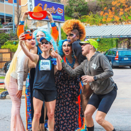 A group of drag queens cheer and pose with two runners taking a selfie on a city street, capturing a lively moment for photography or media requests. One runner holds up a red hat as everyone smiles near a Culver’s sign in the background. Austin Marathon Half Marathon & 5K A group of drag queens cheer and pose with two runners taking a selfie on a city street, capturing a lively moment for photography or media requests. One runner holds up a red hat as everyone smiles near a Culver’s sign in the background. Austin Marathon Half Marathon & 5K