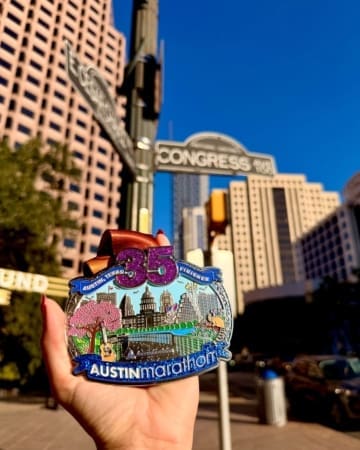 A hand proudly displays marathon medals in front of Congress Ave street signs, with tall downtown buildings and a clear blue sky framing the perfect backdrop for this Austin Marathon finisher medal moment. Austin Marathon Half Marathon & 5K A hand proudly displays marathon medals in front of Congress Ave street signs, with tall downtown buildings and a clear blue sky framing the perfect backdrop for this Austin Marathon finisher medal moment. Austin Marathon Half Marathon & 5K