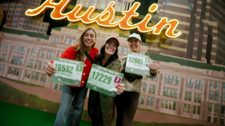 Three smiling women stand in front of a neon "Austin" sign, holding up their race bibs and wearing casual jackets and hats, celebrating together at the Austin Marathon Expo while checking out the latest running gear. Austin Marathon Half Marathon & 5K Three smiling women stand in front of a neon "Austin" sign, holding up their race bibs and wearing casual jackets and hats, celebrating together at the Austin Marathon Expo while checking out the latest running gear. Austin Marathon Half Marathon & 5K