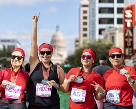 Four women from non profit teams in matching red outfits and medals smile after a race, proudly holding up their race bibs. One woman raises her arm victoriously with the Texas State Capitol visible in the background. Austin Marathon Half Marathon & 5K Four women from non profit teams in matching red outfits and medals smile after a race, proudly holding up their race bibs. One woman raises her arm victoriously with the Texas State Capitol visible in the background. Austin Marathon Half Marathon & 5K