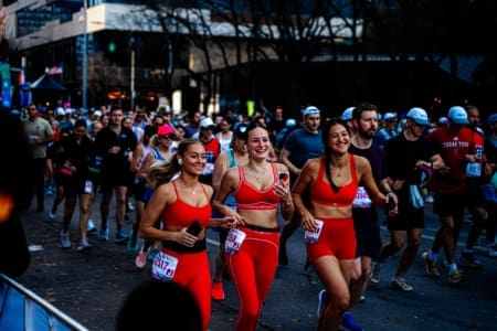 A group of runners participate in a race, with three women in matching bright red outfits—likely part of social teams—smiling and running together in the foreground. Other runners fill the street behind them, showing their dedication to training. Austin Marathon Half Marathon & 5K A group of runners participate in a race, with three women in matching bright red outfits—likely part of social teams—smiling and running together in the foreground. Other runners fill the street behind them, showing their dedication to training. Austin Marathon Half Marathon & 5K