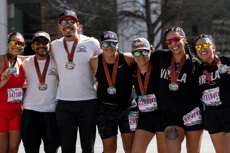 Six smiling runners stand together outdoors, wearing sunglasses, athletic gear, race bibs, and finisher medals around their necks, celebrating as charity teams after completing a race. Austin Marathon Half Marathon & 5K Six smiling runners stand together outdoors, wearing sunglasses, athletic gear, race bibs, and finisher medals around their necks, celebrating as charity teams after completing a race. Austin Marathon Half Marathon & 5K