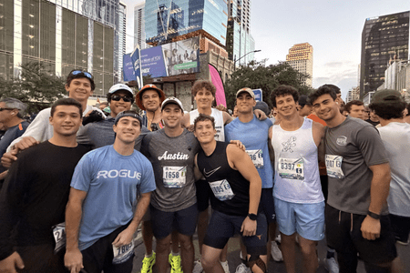 A group of twelve young men from collegiate teams wearing race bibs and athletic clothes smile together at a city street outdoor running event, with tall buildings and trees in the background. Austin Marathon Half Marathon & 5K A group of twelve young men from collegiate teams wearing race bibs and athletic clothes smile together at a city street outdoor running event, with tall buildings and trees in the background. Austin Marathon Half Marathon & 5K