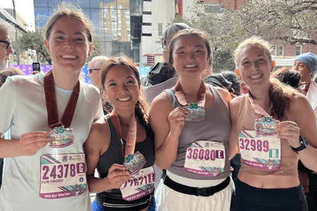 Four smiling women from collegiate teams wearing race bibs and medals pose together after finishing a marathon on a city street. They proudly hold up their medals, dressed in athletic gear, with buildings and trees in the background. Austin Marathon Half Marathon & 5K Four smiling women from collegiate teams wearing race bibs and medals pose together after finishing a marathon on a city street. They proudly hold up their medals, dressed in athletic gear, with buildings and trees in the background. Austin Marathon Half Marathon & 5K