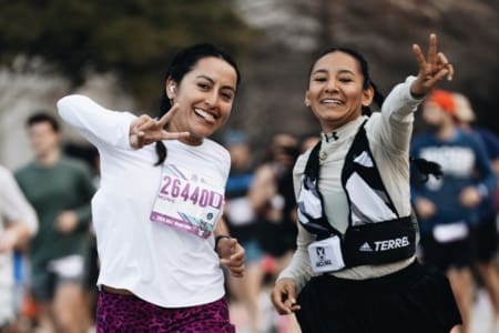 Two women smiling and making peace signs while running in a race as part of community teams, surrounded by other runners. Both wear race bibs and athletic clothing. The background is slightly blurred. Austin Marathon Half Marathon & 5K Two women smiling and making peace signs while running in a race as part of community teams, surrounded by other runners. Both wear race bibs and athletic clothing. The background is slightly blurred. Austin Marathon Half Marathon & 5K