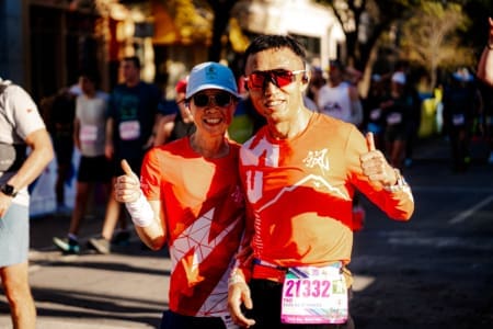 Two smiling runners from the Friends and Family Teams, wearing red shirts, sunglasses, and race bibs, pose with thumbs up at an outdoor race event, surrounded by other participants and trees in the background. Austin Marathon Half Marathon & 5K Two smiling runners from the Friends and Family Teams, wearing red shirts, sunglasses, and race bibs, pose with thumbs up at an outdoor race event, surrounded by other participants and trees in the background. Austin Marathon Half Marathon & 5K