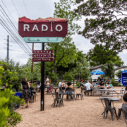 Outdoor patio spot with people sitting at tables under trees. A large vintage "RADIO" sign and smaller signs advertise coffee, cocktails, and live music. It’s a casual, relaxed hangout—perfect for runners enjoying Spring in Austin. Austin Marathon Half Marathon & 5K
