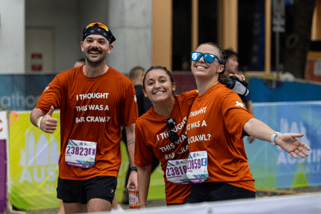 Three smiling runners in matching orange shirts reading "I THOUGHT THIS WAS A BAR CRAWL?" pose happily at a race event, looking like school teams or collegiate teams with their arms around each other and race bibs proudly displayed. Austin Marathon Half Marathon & 5K Three smiling runners in matching orange shirts reading "I THOUGHT THIS WAS A BAR CRAWL?" pose happily at a race event, looking like school teams or collegiate teams with their arms around each other and race bibs proudly displayed. Austin Marathon Half Marathon & 5K