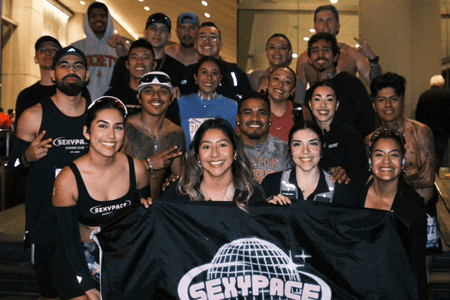 A group of smiling runners from social teams pose together indoors, holding a black "SEXY PACE" flag with a globe graphic. Most wear athletic clothing and race bibs, showing off their training spirit with peace signs and playful gestures. Austin Marathon Half Marathon & 5K A group of smiling runners from social teams pose together indoors, holding a black "SEXY PACE" flag with a globe graphic. Most wear athletic clothing and race bibs, showing off their training spirit with peace signs and playful gestures. Austin Marathon Half Marathon & 5K