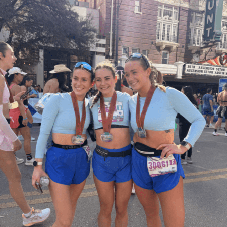 Three women wearing matching light blue crop tops and blue shorts stand together smiling with race bibs and medals, celebrating their achievements in the Austin Marathon team program at an outdoor street event with other participants in the background. Austin Marathon Half Marathon & 5K Three women wearing matching light blue crop tops and blue shorts stand together smiling with race bibs and medals, celebrating their achievements in the Austin Marathon team program at an outdoor street event with other participants in the background. Austin Marathon Half Marathon & 5K