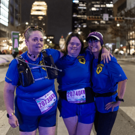 Three women in matching blue shirts and race bibs stand arm-in-arm at a nighttime city race event, representing their company’s Austin Marathon team program, smiling at the camera with tall buildings and bright lights in the background. Austin Marathon Half Marathon & 5K Three women in matching blue shirts and race bibs stand arm-in-arm at a nighttime city race event, representing their company’s Austin Marathon team program, smiling at the camera with tall buildings and bright lights in the background. Austin Marathon Half Marathon & 5K
