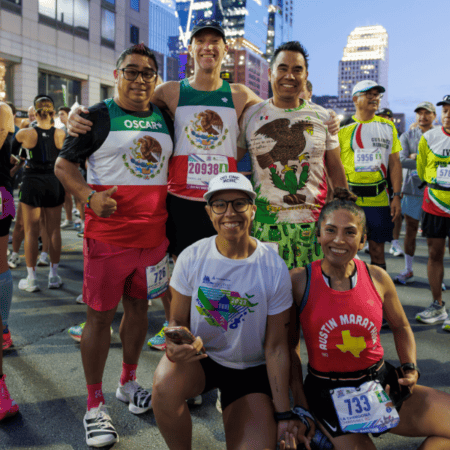 A group of five smiling runners pose together before a race, sporting athletic gear and race bibs. They're participating in the Austin Marathon team program, with a cityscape and fellow Team Registration runners in the background. Austin Marathon Half Marathon & 5K A group of five smiling runners pose together before a race, sporting athletic gear and race bibs. They're participating in the Austin Marathon team program, with a cityscape and fellow Team Registration runners in the background. Austin Marathon Half Marathon & 5K