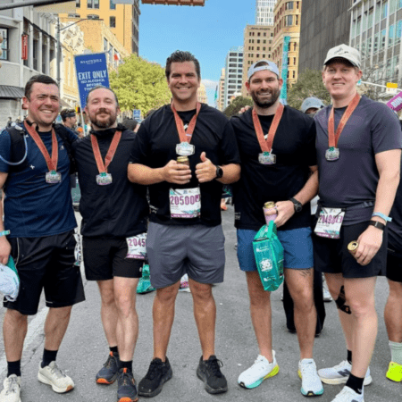 Five men wearing race medals and athletic gear stand together and smile for a photo on a city street after the race, celebrating their Austin Marathon team program success. Buildings and other runners are visible in the background. Austin Marathon Half Marathon & 5K Five men wearing race medals and athletic gear stand together and smile for a photo on a city street after the race, celebrating their Austin Marathon team program success. Buildings and other runners are visible in the background. Austin Marathon Half Marathon & 5K