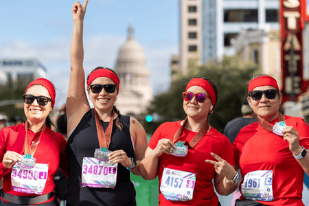 Four women wearing red shirts and headbands proudly display medals after a charity race, smiling for the camera. One woman raises her arm in celebration. City buildings and the Texas State Capitol are visible in the background. Austin Marathon Half Marathon & 5K Four women wearing red shirts and headbands proudly display medals after a charity race, smiling for the camera. One woman raises her arm in celebration. City buildings and the Texas State Capitol are visible in the background. Austin Marathon Half Marathon & 5K
