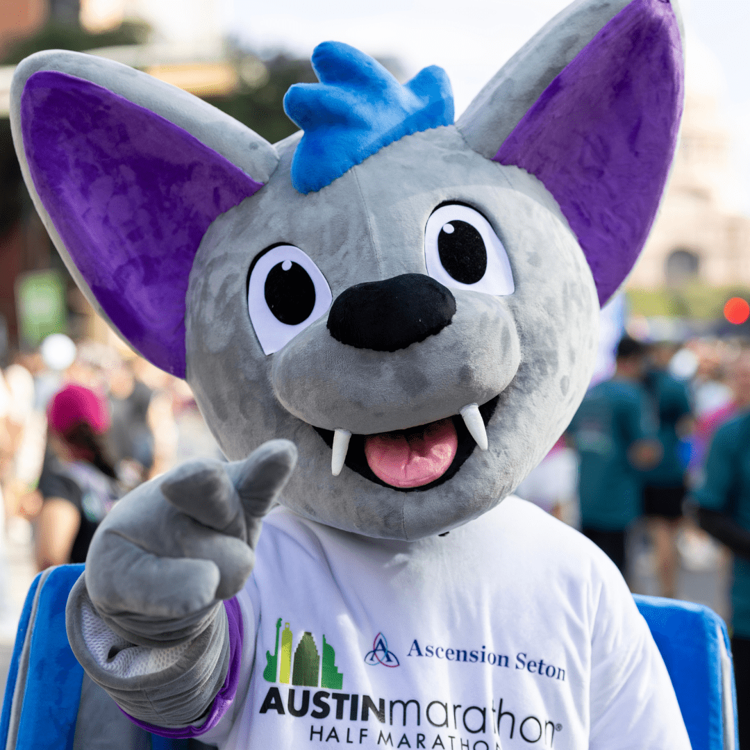 A person in a large, gray bat mascot costume with purple ears and blue accents smiles and points at the camera, proudly sporting an "Ascension Seton Austin Marathon Half Marathon" T-shirt at an outdoor event. Austin Marathon Half Marathon & 5K A person in a large, gray bat mascot costume with purple ears and blue accents smiles and points at the camera, proudly sporting an "Ascension Seton Austin Marathon Half Marathon" T-shirt at an outdoor event. Austin Marathon Half Marathon & 5K