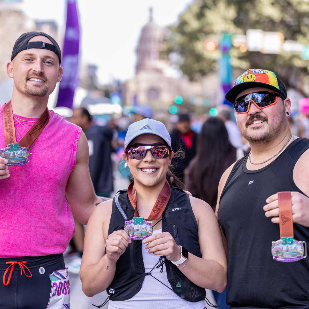 Three runners smile and show off their medals after completing the Ascension Seton Austin Marathon, standing together outdoors with a crowd and city buildings in the background. Austin Marathon Half Marathon & 5K Three runners smile and show off their medals after completing the Ascension Seton Austin Marathon, standing together outdoors with a crowd and city buildings in the background. Austin Marathon Half Marathon & 5K