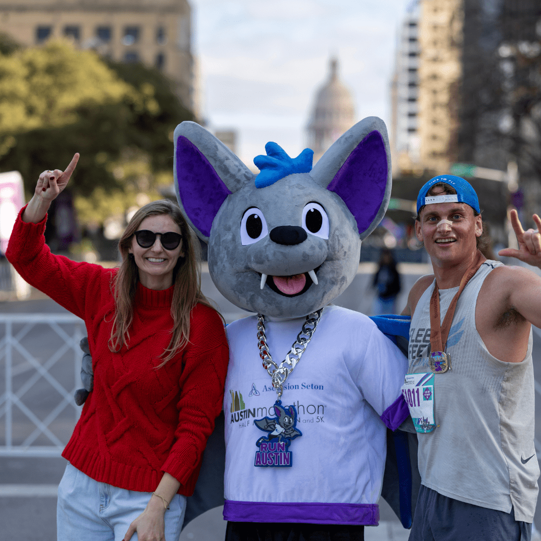 A woman in a red sweater and a man in athletic wear pose with a large bat mascot at an outdoor event, celebrating Austin Marathon history. All three are smiling and making a "number one" gesture against the cityscape and domed building backdrop. Austin Marathon Half Marathon & 5K A woman in a red sweater and a man in athletic wear pose with a large bat mascot at an outdoor event, celebrating Austin Marathon history. All three are smiling and making a "number one" gesture against the cityscape and domed building backdrop. Austin Marathon Half Marathon & 5K