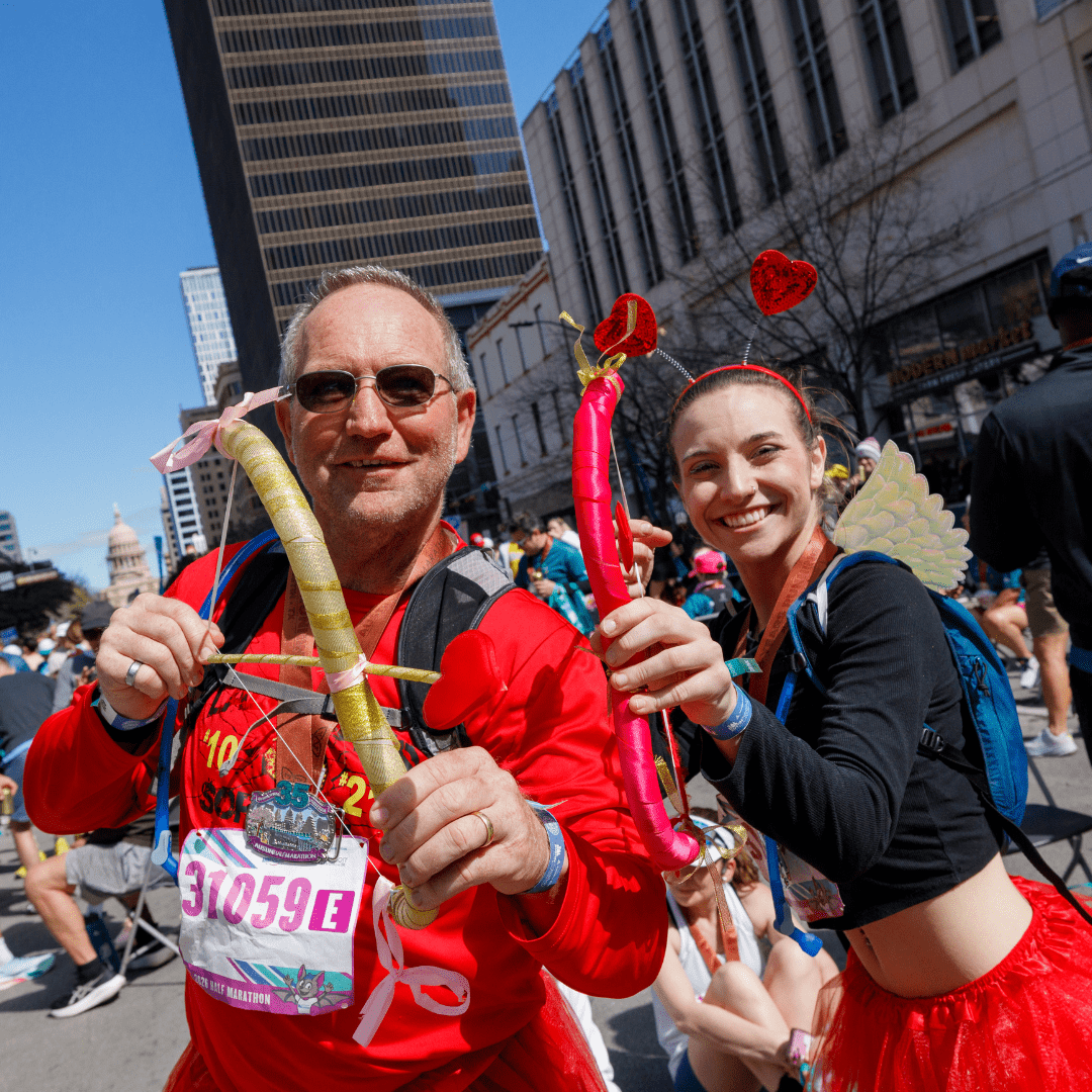 Two people in red costumes with heart-themed accessories and balloon swords smile at a street event, with crowds, tall buildings, and the Texas State Capitol in the background—capturing the festive spirit of Austin Marathon registration day. Austin Marathon Half Marathon & 5K Two people in red costumes with heart-themed accessories and balloon swords smile at a street event, with crowds, tall buildings, and the Texas State Capitol in the background—capturing the festive spirit of Austin Marathon registration day. Austin Marathon Half Marathon & 5K