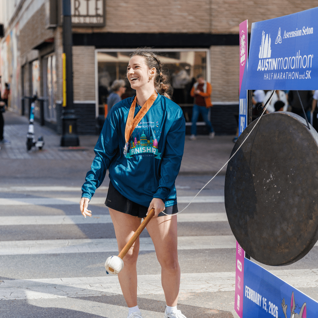 A smiling woman wearing a race finisher medal and shirt stands at the Ascension Seton Austin Marathon finish line, holding a mallet and about to strike a large gong. She appears happy and triumphant. Austin Marathon Half Marathon & 5K A smiling woman wearing a race finisher medal and shirt stands at the Ascension Seton Austin Marathon finish line, holding a mallet and about to strike a large gong. She appears happy and triumphant. Austin Marathon Half Marathon & 5K
