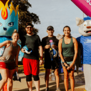A group of five runners from Team Template pose and smile with two mascots at a race event. The inflatable finish line arch behind them reads “Run Austin.” The atmosphere is sunny and festive. Austin Marathon Half Marathon & 5K