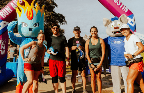 A group of five runners from Team Template pose and smile with two mascots at a race event. The inflatable finish line arch behind them reads “Run Austin.” The atmosphere is sunny and festive. Austin Marathon Half Marathon & 5K