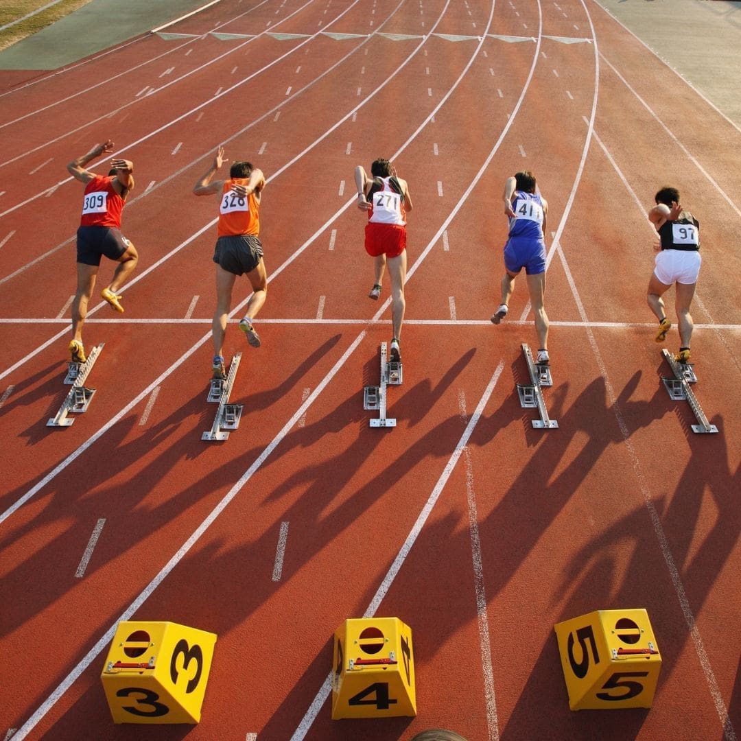 Five sprinters, each in different colored uniforms, launch off starting blocks on a red running track, beginning a race. Yellow lane markers numbered 3 to 7 are visible—reminding first-time marathoners to avoid common marathon mistakes. Austin Marathon Half Marathon & 5K