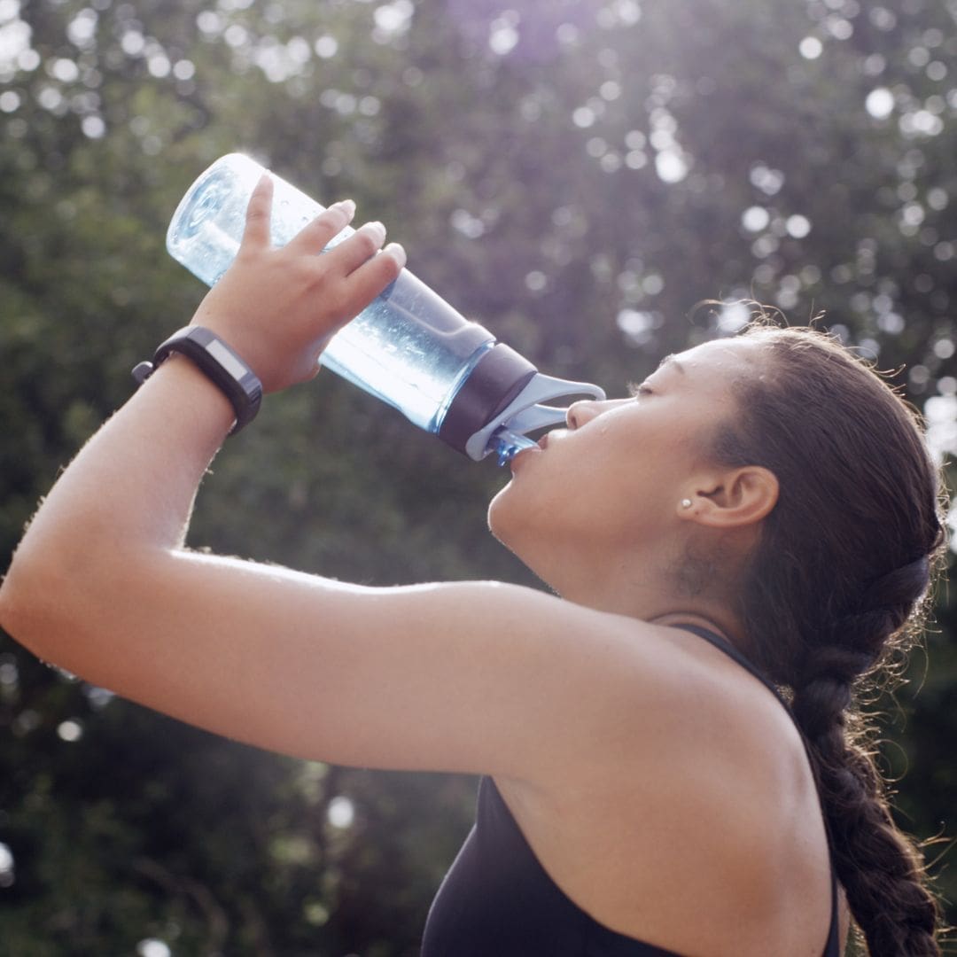 A woman with a braid drinks water from a blue bottle outdoors, wearing a fitness tracker and athletic clothing—highlighting essential marathon tips for first-time marathoners as sunlight filters through the trees in the background. Austin Marathon Half Marathon & 5K