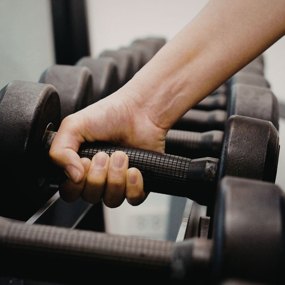 A close-up of a hand gripping the handle of a black dumbbell, with several others in a rack behind—perfect for first-time marathoners looking to avoid common marathon mistakes by adding strength training to their routine. Austin Marathon Half Marathon & 5K