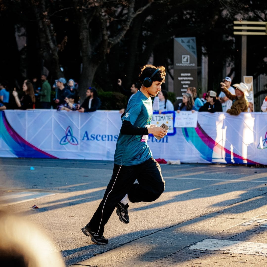 A runner wearing headphones and a teal shirt jogs past cheering spectators and a white event banner on a sunny day, embodying the excitement and energy first-time marathoners feel while learning valuable marathon tips along the route. Austin Marathon Half Marathon & 5K