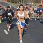 A woman wearing a white sports bra and blue shorts runs in a road race, holding a baton. Other runners follow behind her on a sunlit street, showcasing the spirit of summer running. Austin Marathon Half Marathon & 5K