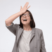 A woman wearing glasses and a gray blazer smiles with her eyes closed, touching her forehead in a classic "facepalm" gesture, capturing the patience needed for marathon training or the underrated skill of laughing at your own slip-ups. Austin Marathon Half Marathon & 5K