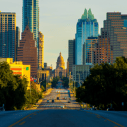 A view of downtown Austin, Texas, with the Texas State Capitol framed by skyscrapers—an iconic scene along an Austin bucket-list marathon route, seen from a wide tree-lined street in the glow of early morning sunlight. Austin Marathon Half Marathon & 5K