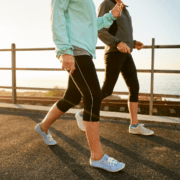 Two people in athletic wear power walk on a paved ocean path, sunlight behind them. Only their legs and torsos are visible—showing a smart way to build running distance and avoid burnout while enjoying scenic exercise. Austin Marathon Half Marathon & 5K