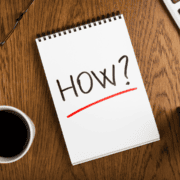 A spiral notebook on a wooden desk displays the word “HOW?” in bold black letters with a red underline. A cup of coffee and part of a keyboard are visible nearby, hinting at February plans for endurance or heat training sessions ahead. Austin Marathon Half Marathon & 5K