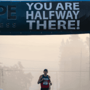 A runner in a race passes under a large banner that reads "YOU ARE HALFWAY THERE!"—a perfect scene for a February Goal or Mid-Year Fitness Check, with fog and trees visible in the background. Austin Marathon Half Marathon & 5K
