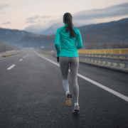 A woman wearing a teal jacket and gray leggings runs alone on an empty highway with mountain scenery in the background under a cloudy sky, staying focused on her February goal. Austin Marathon Half Marathon & 5K
